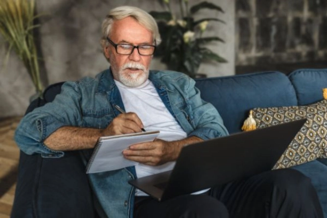 Older man on a sofa taking notes beside a laptop