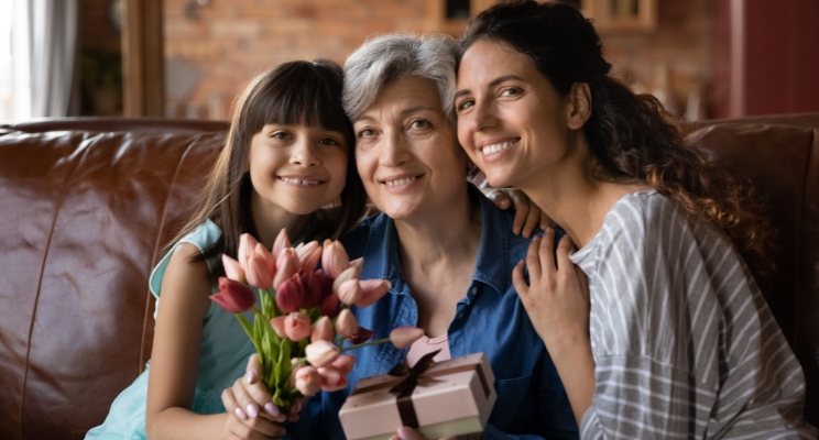 Grandmother holding flowers and a gift, hugged by her daughter and granddaughter